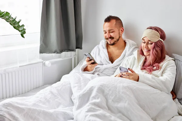 Casal caucasiano usando smartphone deitado na cama juntos, verificando redes sociais notícias — Fotografia de Stock