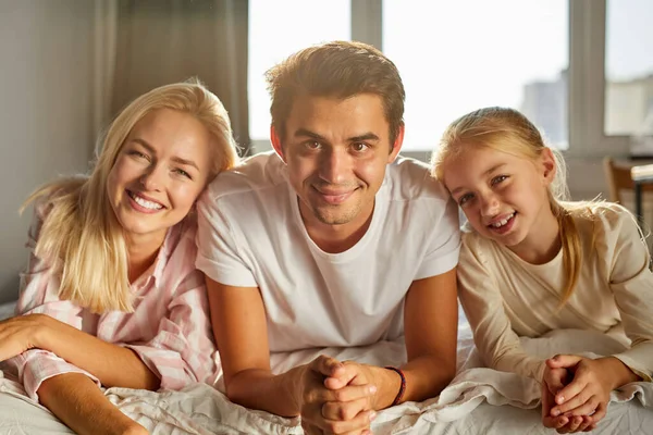 Belle famille caucasienne composée de femme homme et enfant sourire à la caméra couché sur le lit — Photo