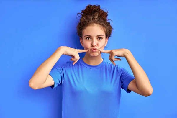 Διασκέδαση έφηβος γυναίκα δείχνει δείκτη δάχτυλα για φυσώντας μάγουλα που απομονώνονται στο μπλε — Φωτογραφία Αρχείου