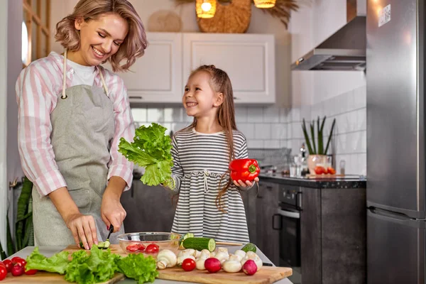 Attraktive junge alleinerziehende Mutter, die mit ihrer Tochter in der Küche einen Salat zubereitet — Stockfoto