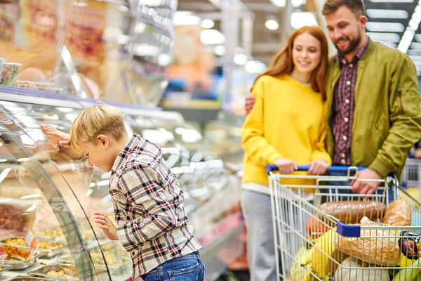 Kleine jongen leunde op showcase met voedsel in petto, ouders op de achtergrond — Stockfoto