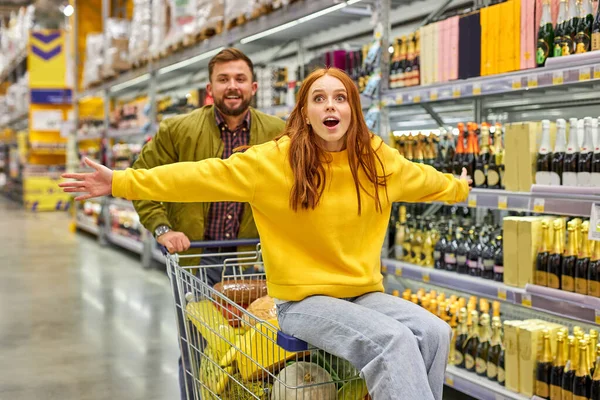 man pushes shopping cart with redhedad woman sitting in it