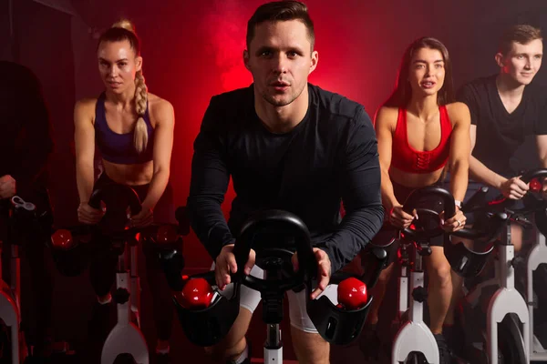 Muscular jovem caucasiano homem exercitando cardio treinamento em bicicleta em fitness ginásio com amigos — Fotografia de Stock