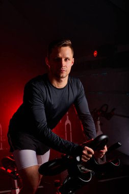 Spor salonunda bisiklete binip kameraya poz veren yakışıklı, beyaz bir erkek.