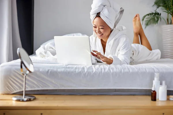 Positive asian woman in white bathrobe in bedroom at home typing message to friends using laptop — Stock Photo, Image