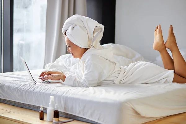 Позитивная азиатка в белом халате в спальне дома, печатает сообщения друзьям с помощью ноутбука — стоковое фото