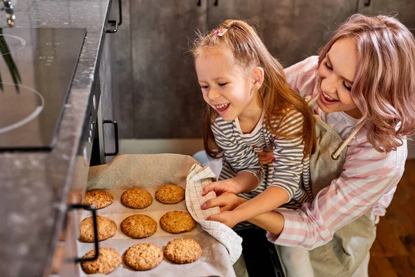 Szczęśliwa córka i matka wyciągając tacę pieczonych ciasteczek z piekarnika w kuchni — Zdjęcie stockowe