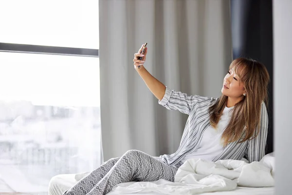 Молодая азиатка в пижаме делает селфи на смартфоне — стоковое фото