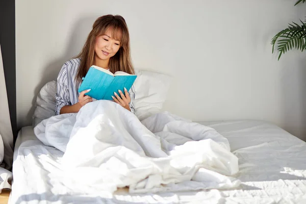 Bonita asiático mulher senta leitura livro no cama — Fotografia de Stock