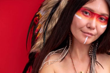 Kafasında tüyle gülümseyen şaman kadın kameraya bakıp gülümsüyor.