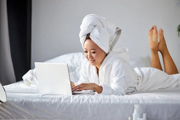 Attractive woman in towel and bathrobe lying on bed at home and working on her laptop — Stock Photo, Image