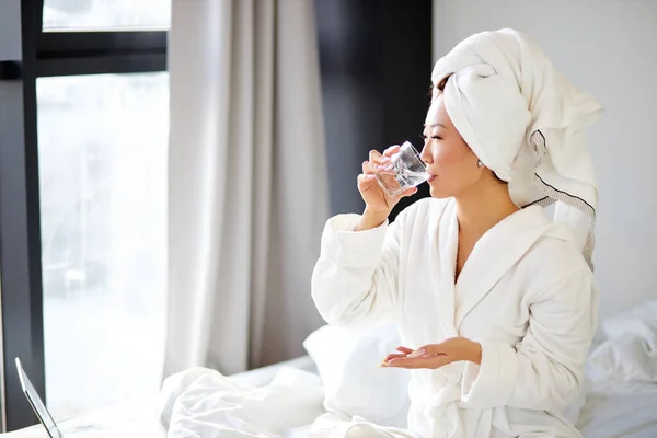 Chinoise prenant une pilule, assise sur son lit à la maison, portant peignoir et serviette — Photo