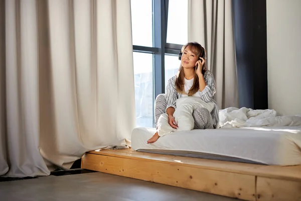 Adorable asiático hembra disfruta de música en auriculares sentado en cama — Foto de Stock