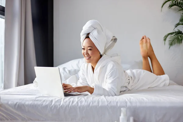 Vacker kvinna i handduk och badrock ligger på sängen hemma och arbetar på sin bärbara dator — Stockfoto