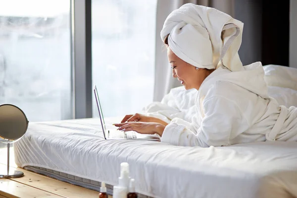 Hermosa mujer en toalla y albornoz acostada en la cama en casa y trabajando en su computadora portátil — Foto de Stock