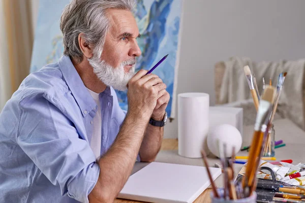 Portrait of mature artist sitting behind table and thinking — Stock Photo, Image