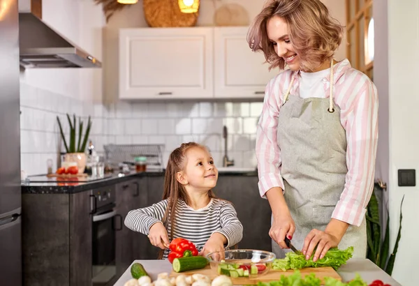 Jolly child girl preparing a salad with her mother in the kitchen — Stock Photo, Image