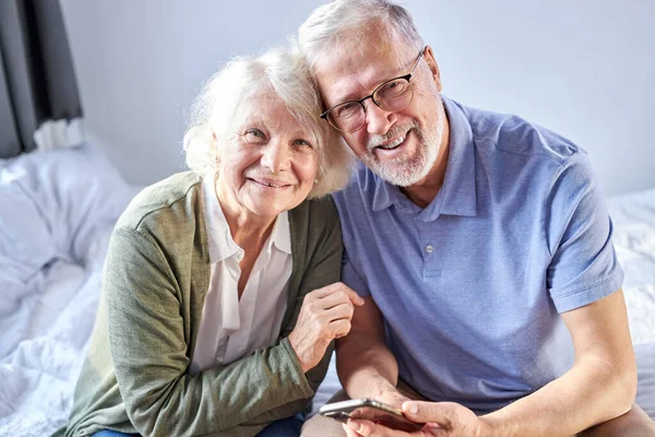 Retrato de idosos sorridentes 60 anos marido e mulher sentar com smartphone relaxar na cama abraçando — Fotografia de Stock