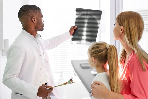African male pediatrician explaining and showing lung x-ray for sick child and mother in hospital office — Foto Stock