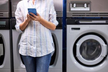 Çamaşır yıkamanın tadını çıkarıyorum. Çamaşır makinesinin yanında duran ve akıllı telefon uygulamasına bakan bir kadının kesilmiş fotoğrafı.