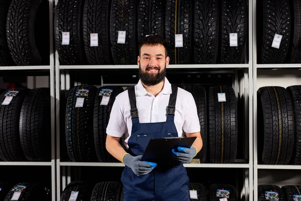 Giovane meccanico maschio caucasico con appunti vicino agli pneumatici nel centro di assistenza automobilistica — Foto Stock