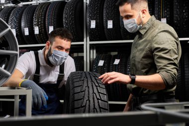 Tıbbi maskedeki satıcı ve müşteri araba lastiklerini tartışıyor.