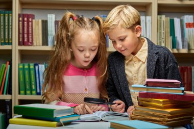 Okul çocukları kütüphanede masada otururken birlikte kitap okuyorlar.