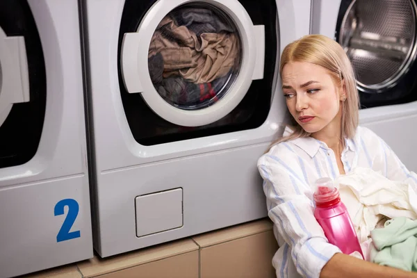 Красивая молодая надоедливая женщина сидит у стиральной машины, ожидая окончания стирки — стоковое фото