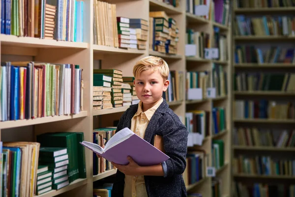 portrait of diligent caucasian child boy with book between bookshelves in campus library