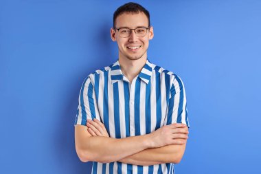 Çizgili tişörtlü, gözlüklü, kolları katlanmış ve gülümseyen neşeli bir erkek.