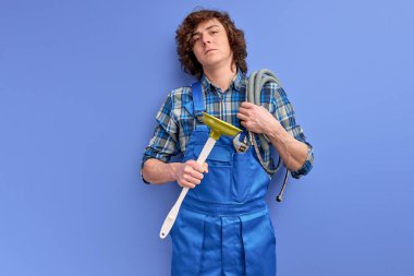 Üniformalı, kıvırcık saçlı, kendine güvenen tesisatçı mavi arka planda tuvalet pompası tutuyor.