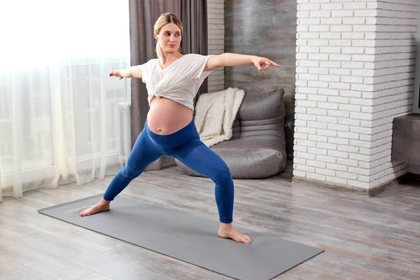 Giovane donna incinta a piedi nudi flessibile in leggins e t-shirt stretching gambe mentre fa esercizi di yoga — Foto Stock