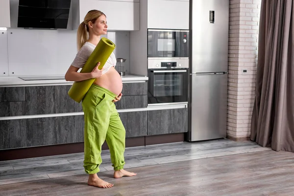 Vista lateral de la mujer embarazada con gran barriga mirando hacia un lado, sosteniendo la esterilla de fitness en las manos —  Fotos de Stock