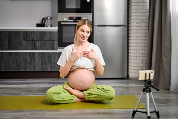 Dost canlısı hamile kadın spor paspasına oturup akıllı telefondan video kaydediyor. — Stok fotoğraf