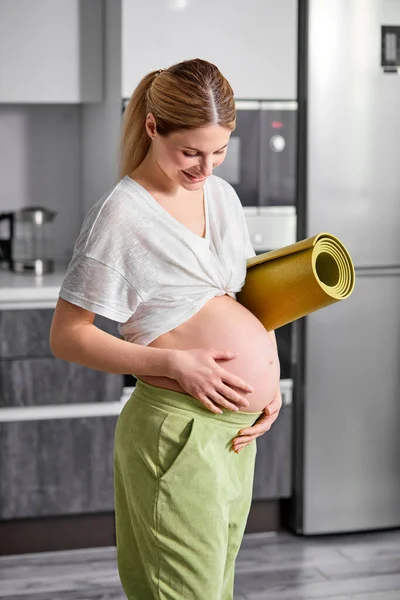 Retrato de la mujer embarazada en camiseta blanca y pantalones verdes de pie, abrazando la barriga — Foto de Stock