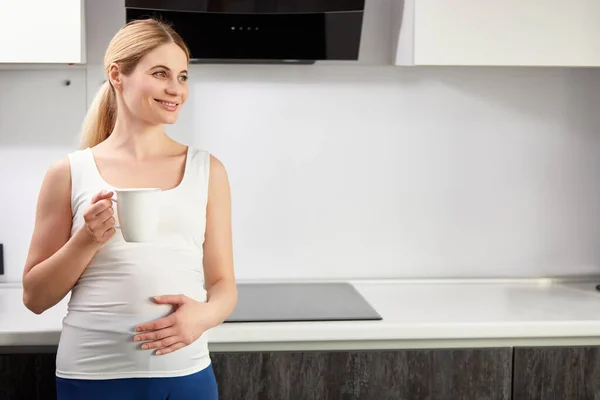 Hermosa mujer embarazada feliz beber taza de té o café en la cocina en casa, por la mañana —  Fotos de Stock