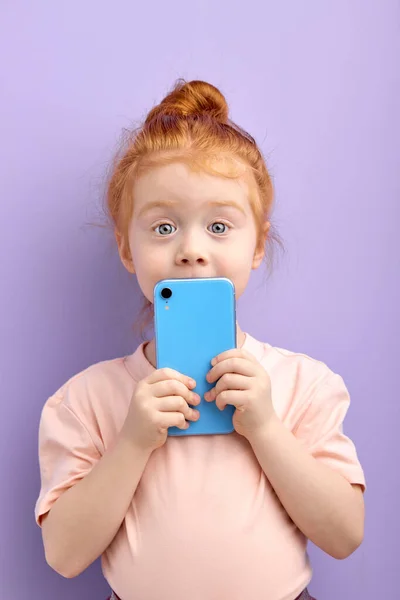 스마트폰을 사용하는 예쁜 빨간 머리 여자 아이의 모습, 스튜디오의 보라색 배경에 고립된 모습. — 스톡 사진