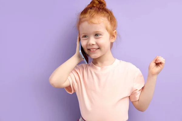 En liten rödhårig tjej som pratar i telefon inomhus. begreppet beroende av barn och prylar — Stockfoto