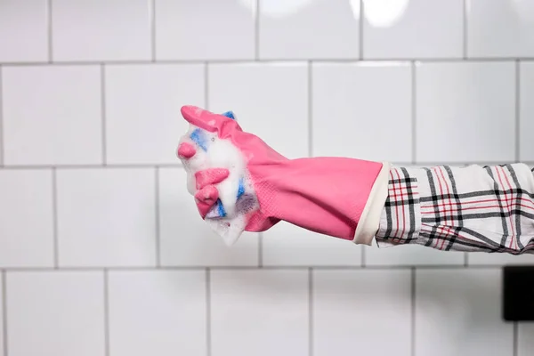 Guante de mano en goma que sostiene la esponja en espuma, aislado en la pared de azulejos blancos — Foto de Stock