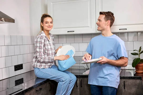 Agradable mujer caucásica limpiando los platos mientras el marido se lava en la cocina —  Fotos de Stock