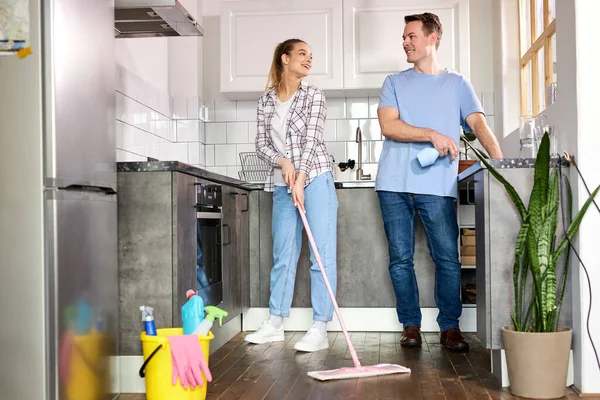 Amichevole giovane coppia pulizia casa, in cucina. Donna che pulisce il pavimento con la scopa, marito che aiuta — Foto Stock