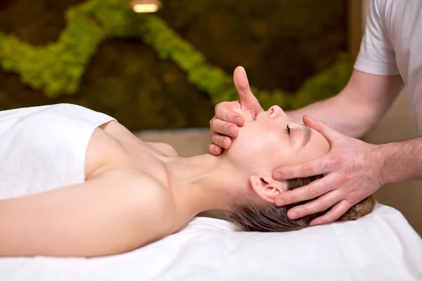 Joven hermosa mujer disfrutando de masaje facial. Hombre terapeuta haciendo masaje de cabeza y cuello a cliente femenino, vista lateral — Foto de Stock