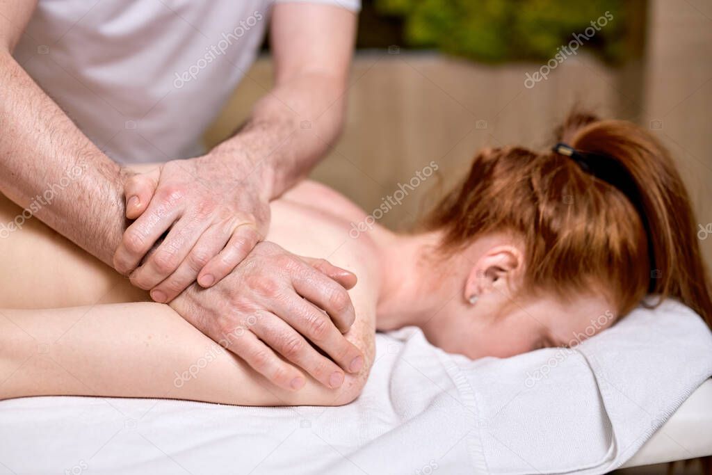 Side View on unrecognizable redhead woman enjoying massage on back and shoulders lying on belly.