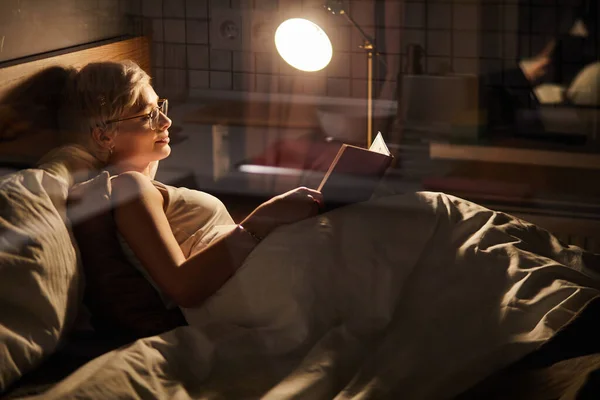 Femmina in occhiali sdraiata sul letto lettura libro interessante preferito a casa in camera illuminata da lampada — Foto Stock