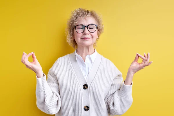 Starší žena v brýlích zachovat klid, namaste, izolované přes žluté pozadí — Stock fotografie