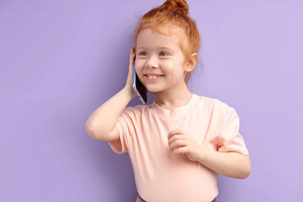 Caucasico rossa ragazza bambino comunica sul telefono cellulare, sorride e parla isolato su sfondo viola — Foto Stock