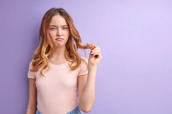 Δυσαρεστημένο νεαρό καυκάσιο κορίτσι αγγίζει τα μαλλιά απομονωμένο σε μωβ φόντο στούντιο πορτρέτο — Φωτογραφία Αρχείου