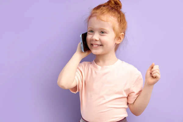 Fiatal gyermek lány kommunikál mobiltelefonon, mosolyog és beszél elszigetelt lila háttér — Stock Fotó