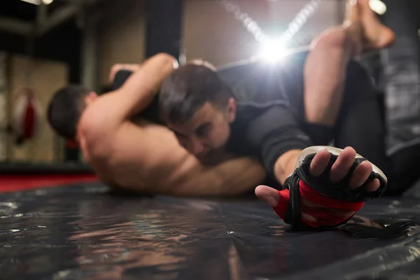 Jóvenes luchadores de MMA luchando contra el entrenamiento en el gimnasio. Deportista musculoso tumbado en el suelo y resistiendo, de cerca las manos —  Fotos de Stock