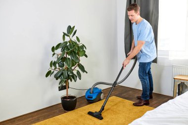 Modern beyaz oturma odasında elektrikli süpürgeyle halı temizleyen yakışıklı beyaz adam.